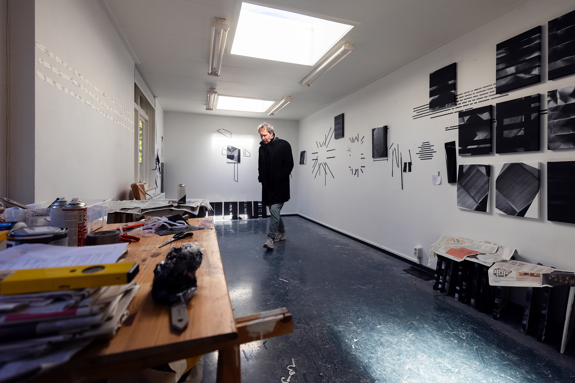 René staat midden in het atelier in een lange zwarte jas, kijkend naar de vloer en omringd door zijn abstracte schilderijen
